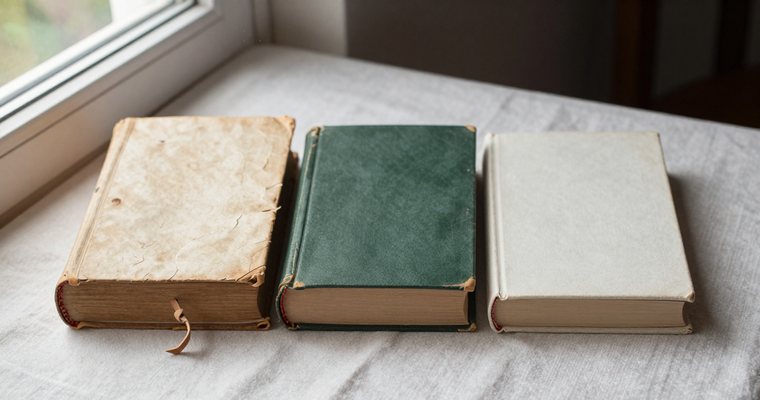 Three books side by side on pale linen: a warped vellum-bound folio, a worn green cloth volume, and a plain modern one